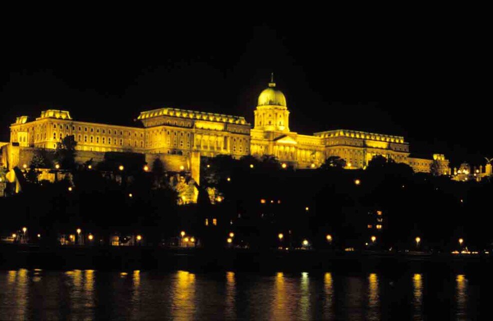 Una foto notturna del grosso edificio del parlamento di budapest si staglia oltre la riva del danubio.