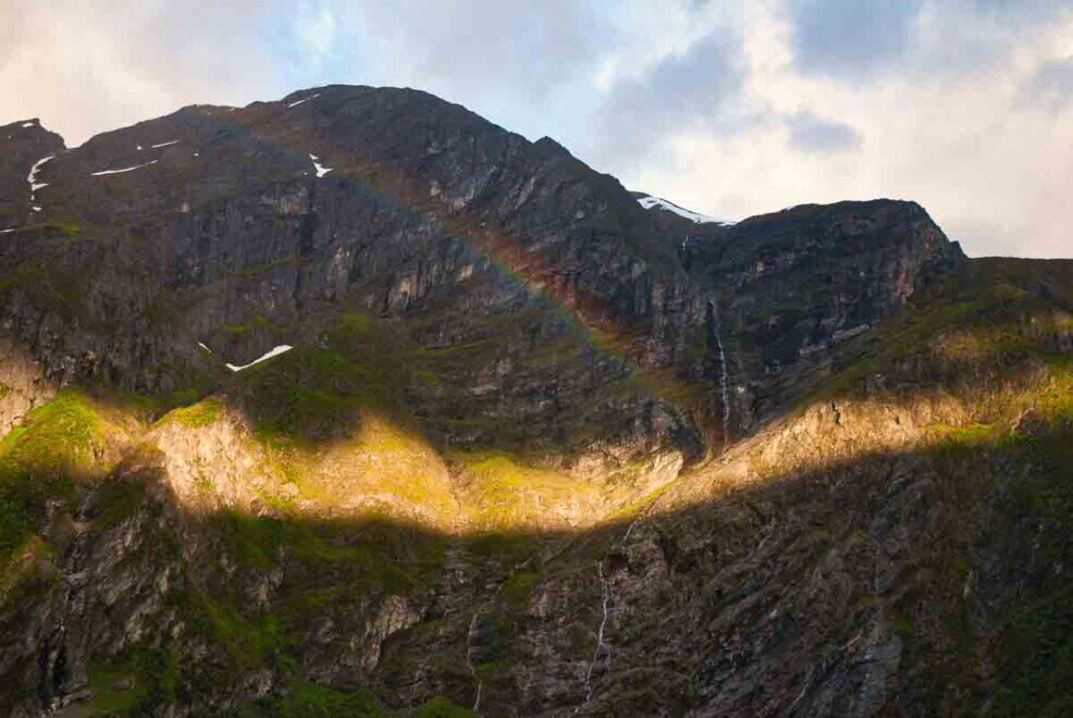 Geiranger Fjord: in uno spiraglio fra le nuvole filtra la luce del sole, e un arcobaleno si riflette sul costone della montagna.