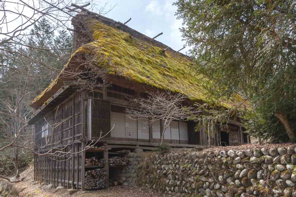 Casa contadina in legno con tetto molto spiovente ricoperto di vegetazione e circondata da un muretto in pietre