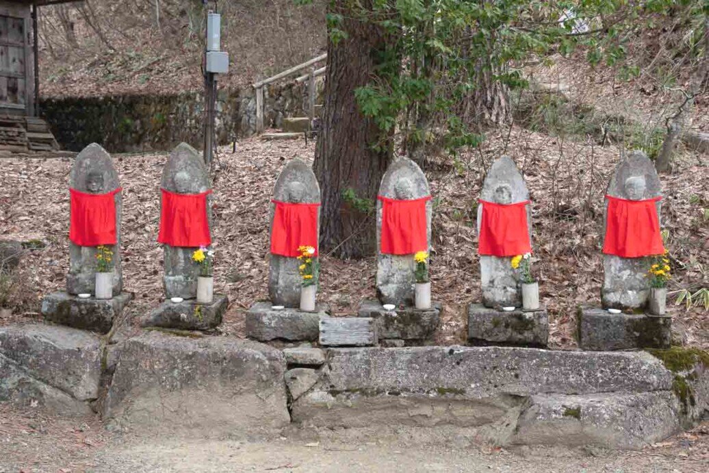 Sei piccole statuine di jizo una a fianco all'altra posizionate all'aperto e ciascuno con un bavaglino rosso.