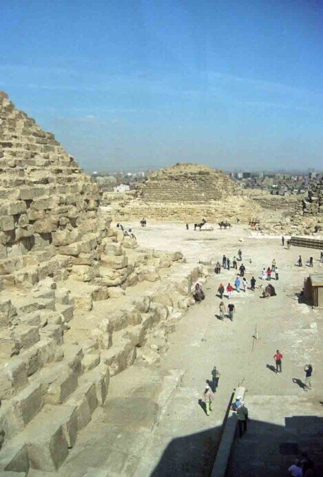 La piramide di Cheope con una parete parzialmente crollata e lo sullo sfondo la città molto vicina.