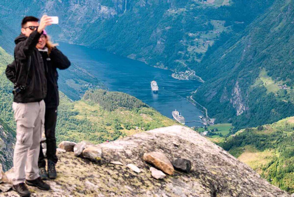 Una coppia si fa un selfie sopra il monte Dasnybba con in basso il porticciolo di Geiranger