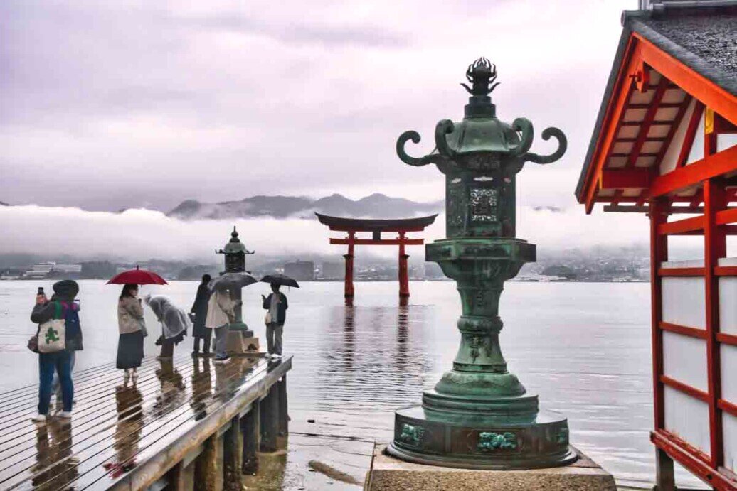 Una lanterna in pietra vicino ad un molo con sullo sfondo un torii rosso con la base immersa nell'acqua