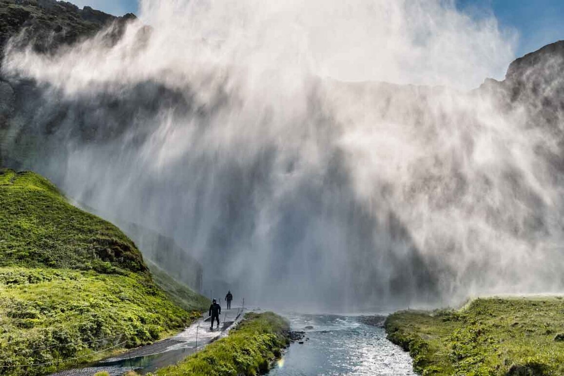 Grande cascata che scende da una parete rocciosa, con persone, più sotto, che camminano lungo un sentiero in primo piano.